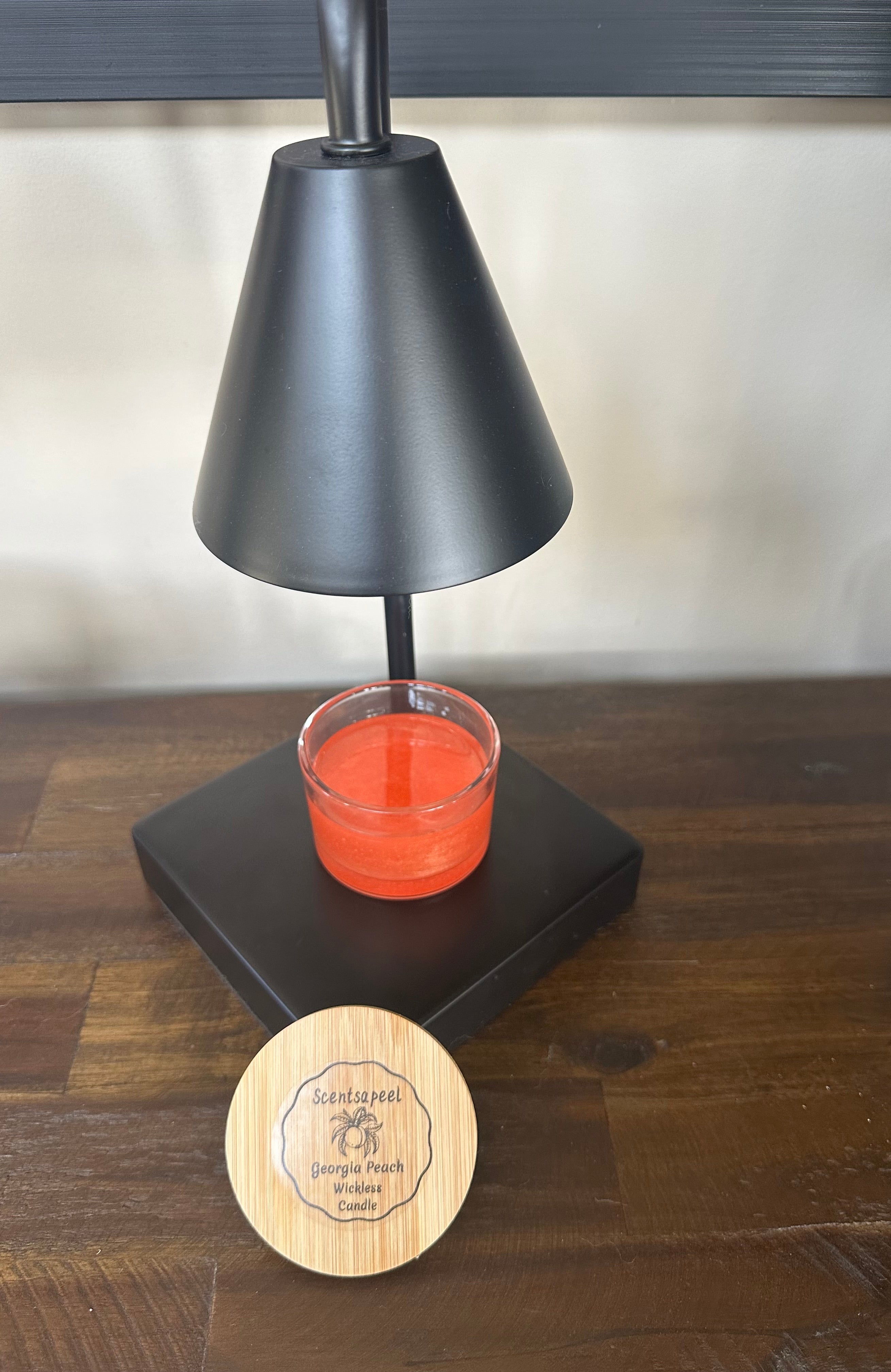 Black candle warming lamp turned off with a peach scented wickless candle sitting on base with wooden lid leaning against base of lamp on a wood dresser