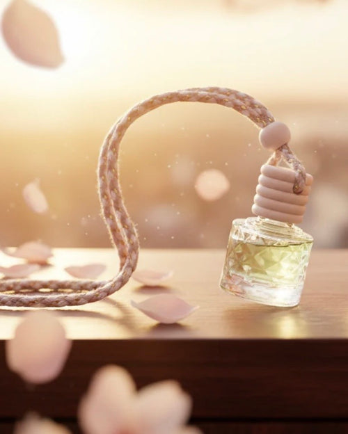 A small clear glass car diffuser bottle filled with fragrance oil attached to a braided beige cord and white beaded top, resting on a wooden surface with soft petals and a warm blurred background.