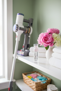 Scentsapeel Cold Air Diffuser Spray on a shelf in a laundry room setting with vacuum leaning on shelf  with flowers and folded towels and a wicker basket